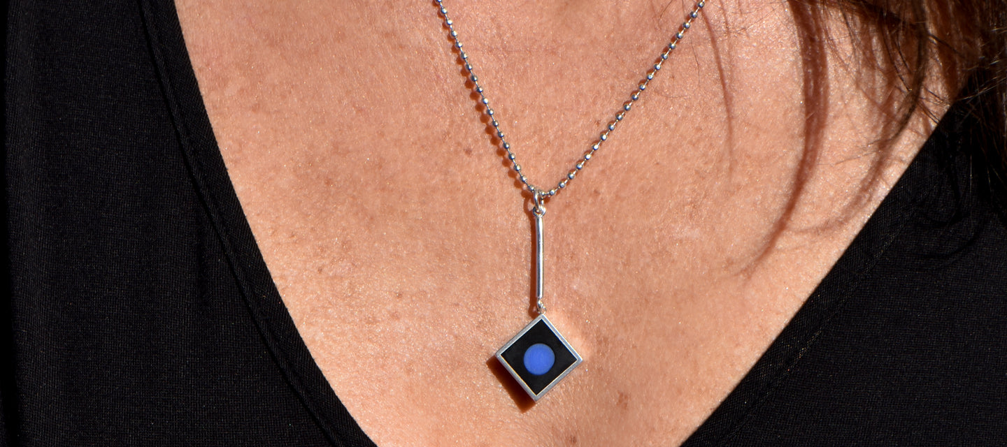 A blue glass dot is set in a sterling silver square pendant, embedded in black resin and hanging from a silver chain.