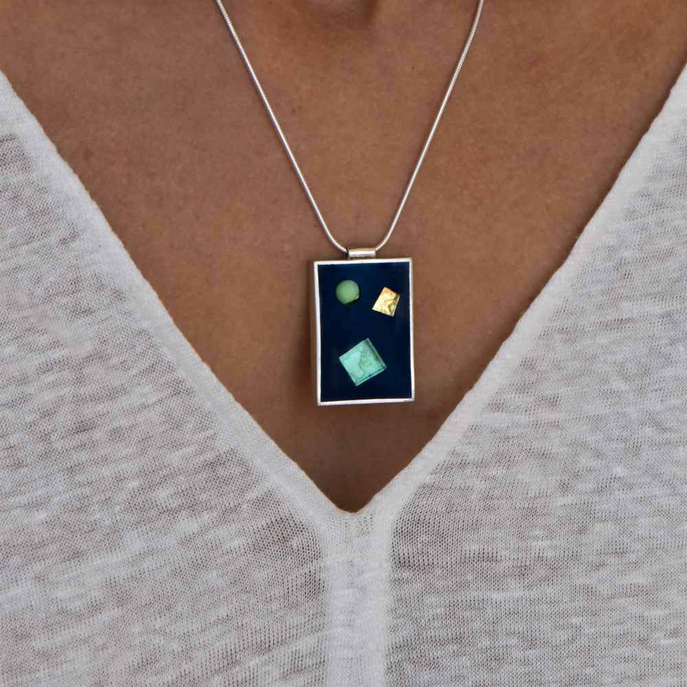 Green and gold-fused glass are embedded in black resin sitting in a rectangular sterling silver setting, hanging from a sterling silver chain.