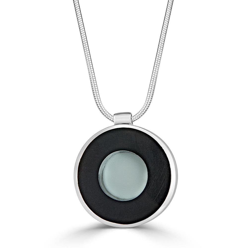 Transparent Beauty Necklace by Ronnie Taubenfeld is a transparent glass disk with a blue-gray backing embedded in black resin and set in a circular silver frame, suspended by a silver snake chain.